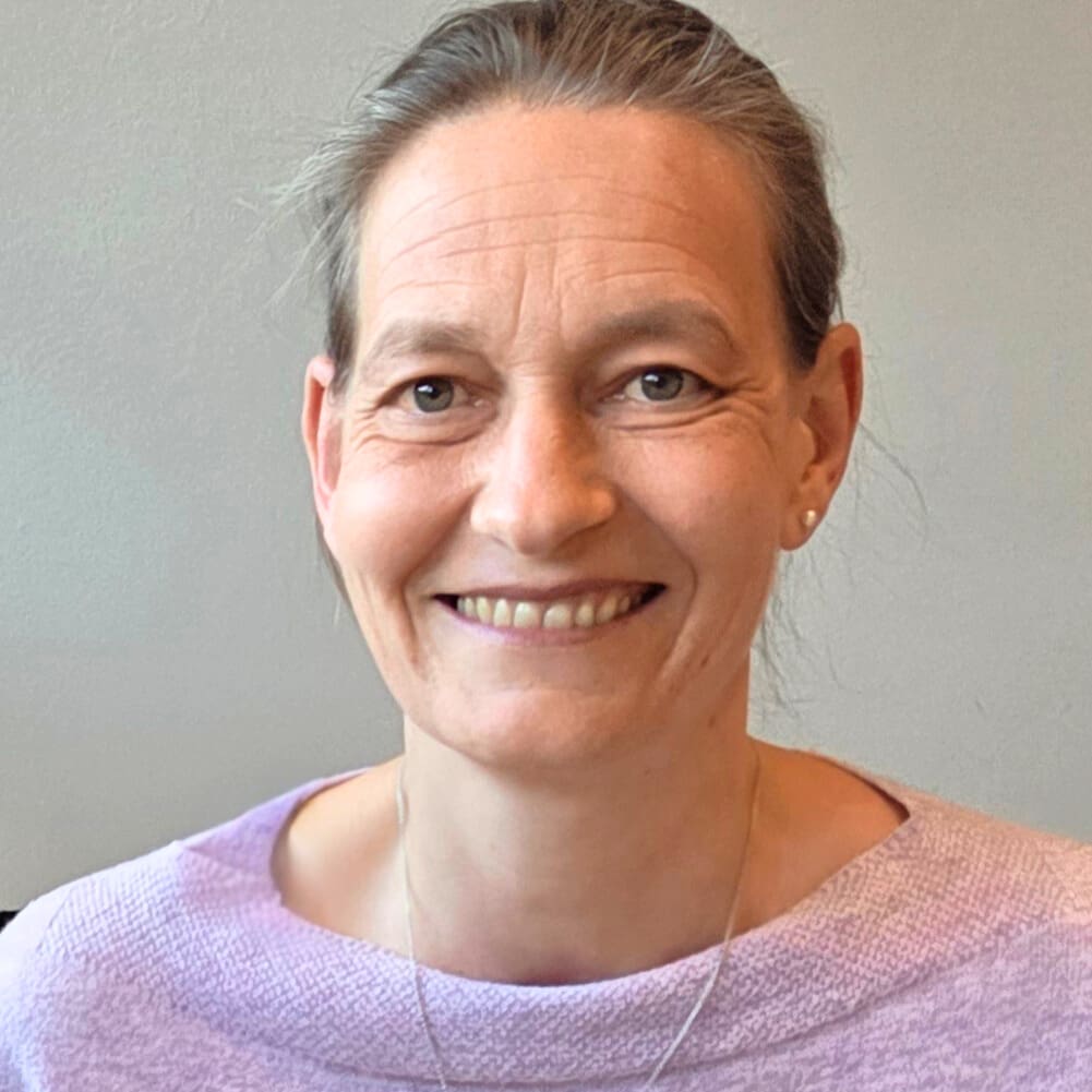 Evigo® eine frau mit heller haut und zurückgebundenen braunen haaren lächelt in die kamera. sie trägt einen hellvioletten pullover und einen kleinen ohrring, bereit, ihr ansprechpartner vor einem schlichten hintergrund zu sein.